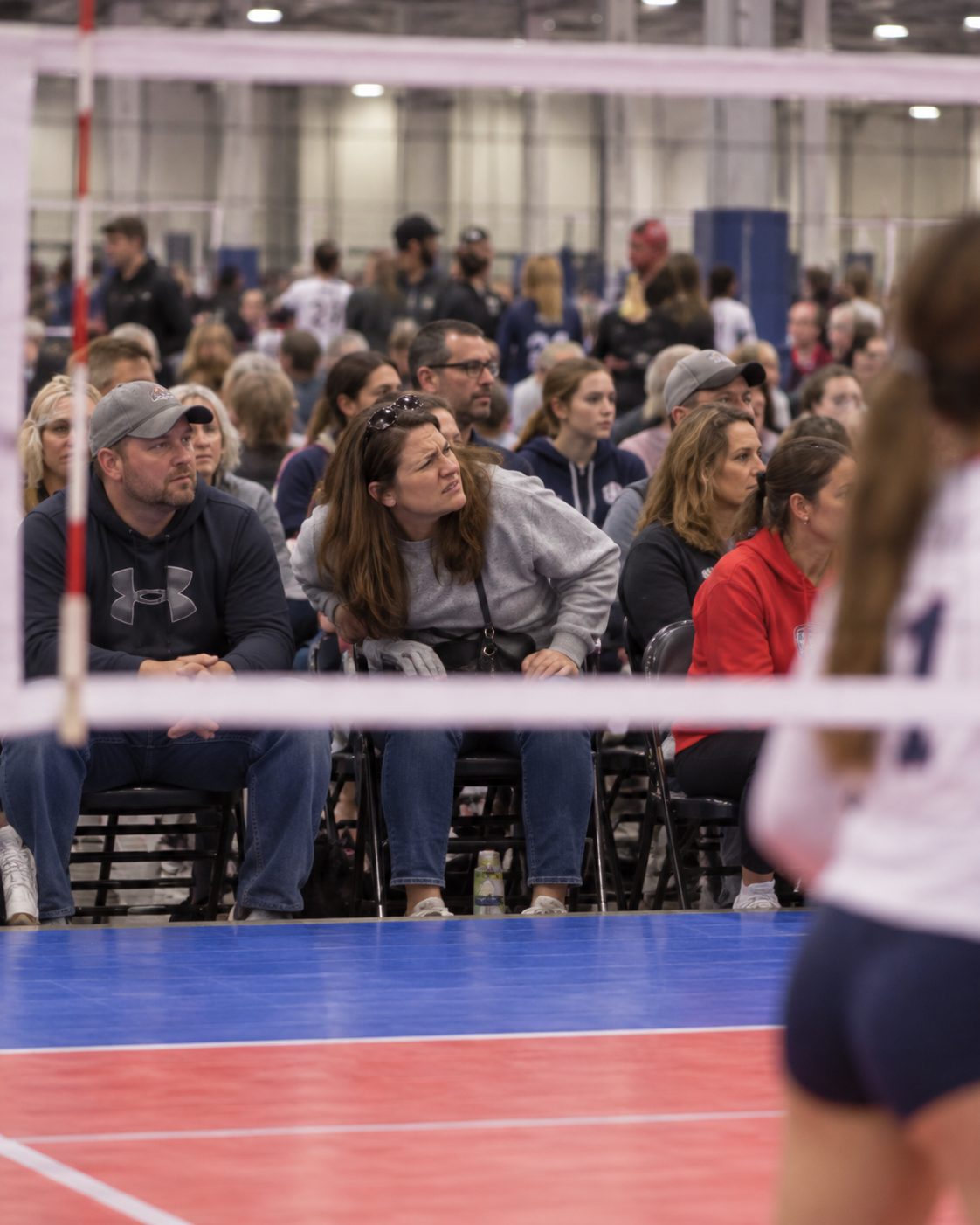 A spectator in the front row leans forward, trying to see past the volleyball net.